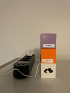 Auf einem Kühlschrank liegt eine schwarze Mehrfachsteckdose, mit der das Smart Home System "homee" verbunden ist. Dieses besteht aus den aufeinander gestapelten weißem homee brain Würfel, dem orangen Zigbee Würfel und dem lila Z-Wave Würfel. Auf dem weißen Würfel ist das Logo von "homee" zu sehen.  